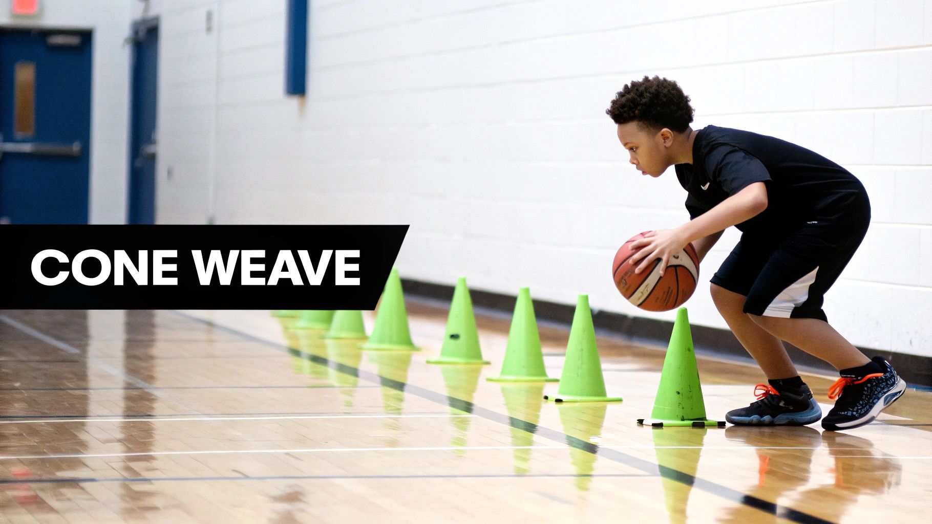 A young boy practices basketball dribbling by weaving through a line of green cones in a gym.