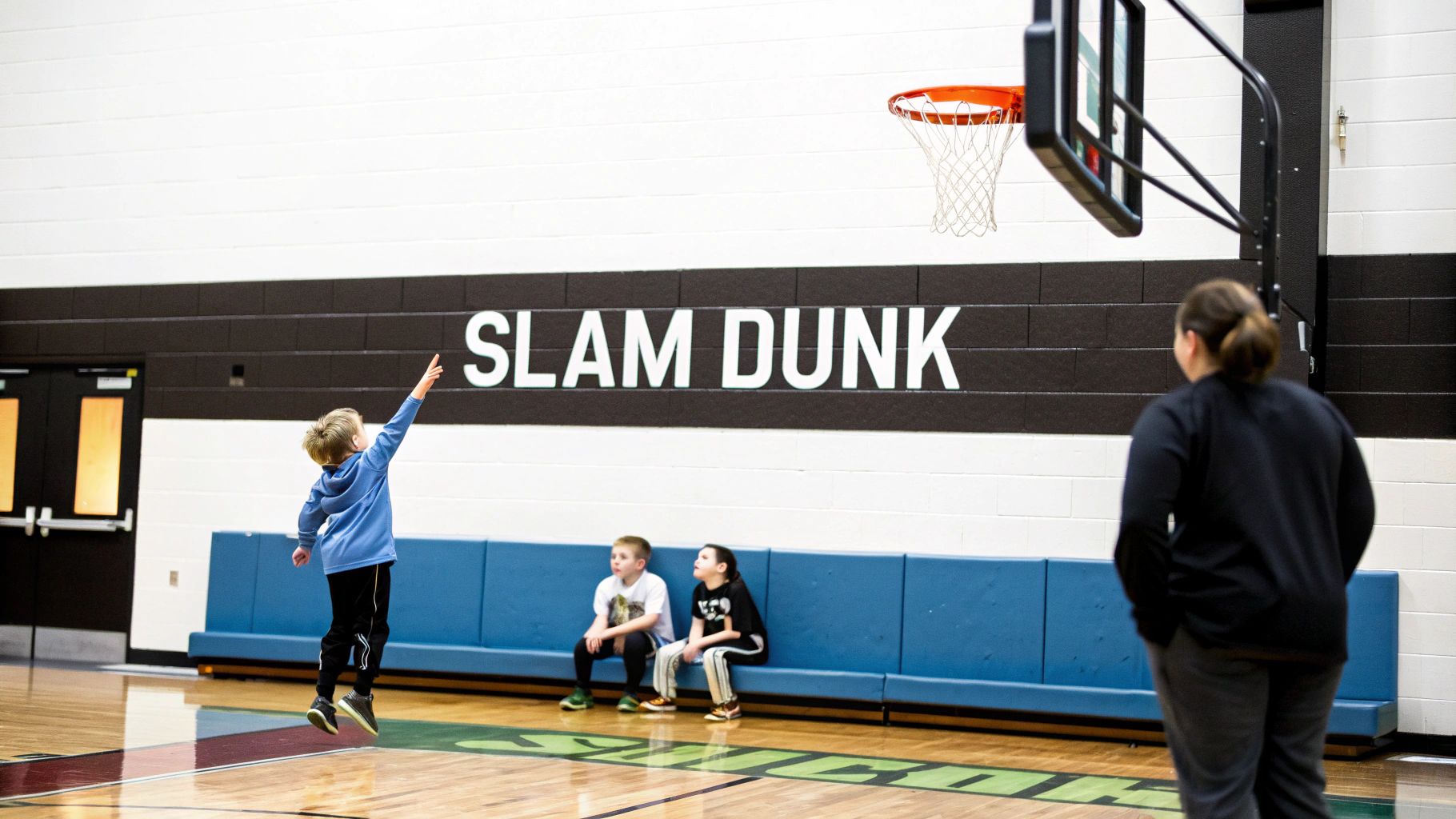 A young boy jumps to reach a basketball hoop on a court with other children and an adult.