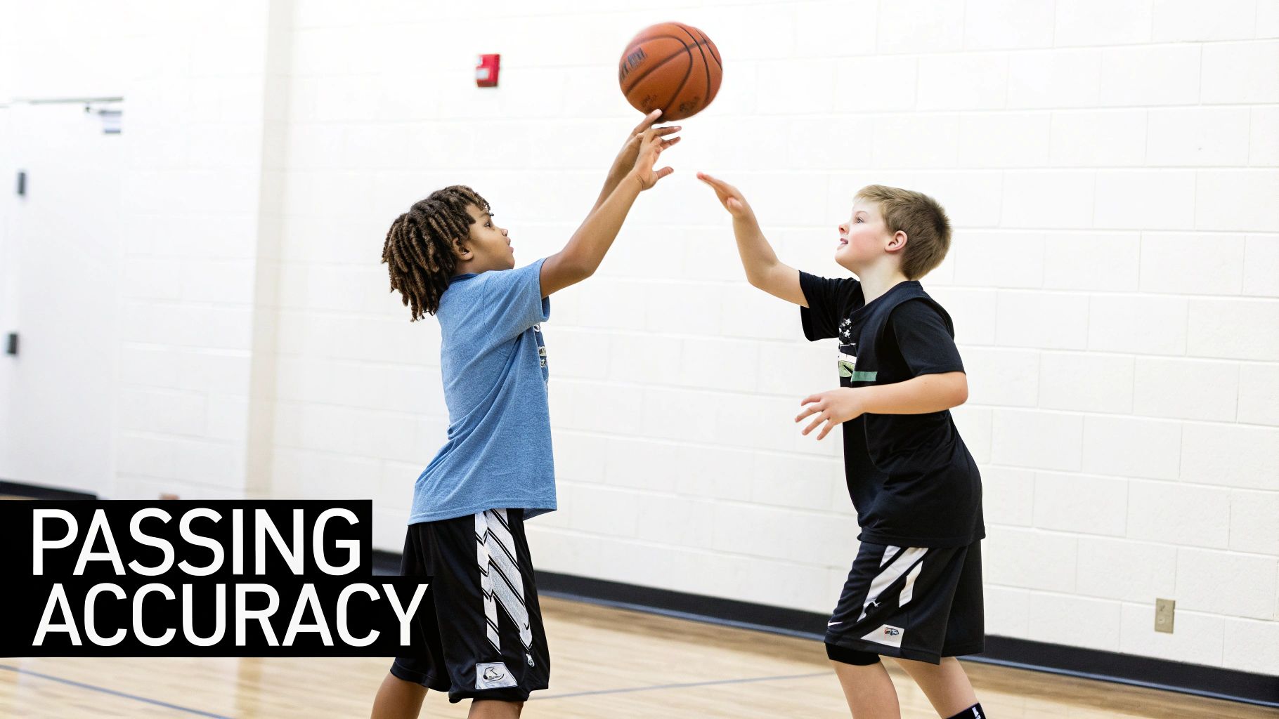 Two young boys in a gym practicing basketball, one passing the ball to the other who reaches to catch it.