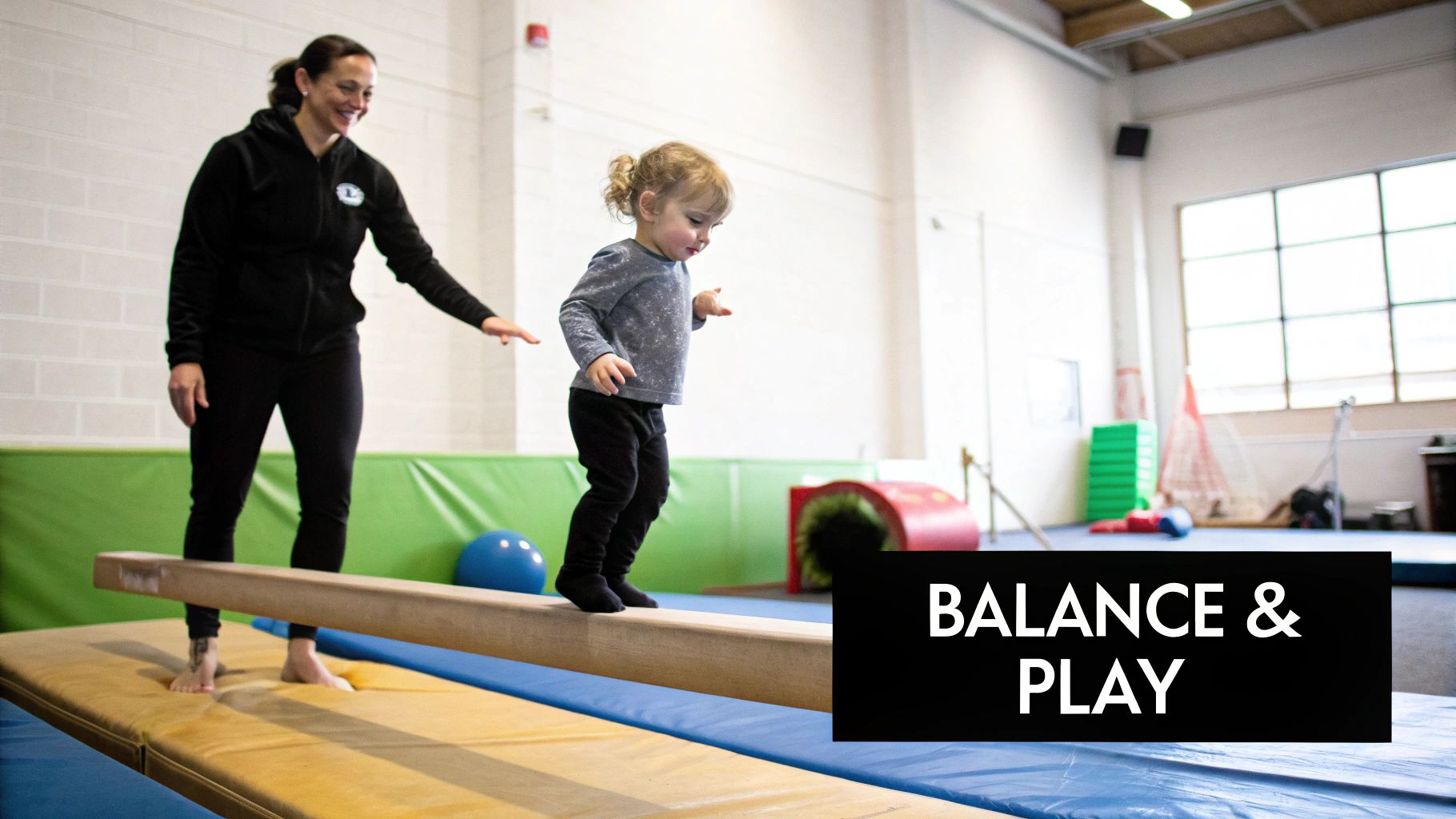 A young child walks on a low balance beam in a gym, supervised by a smiling adult.