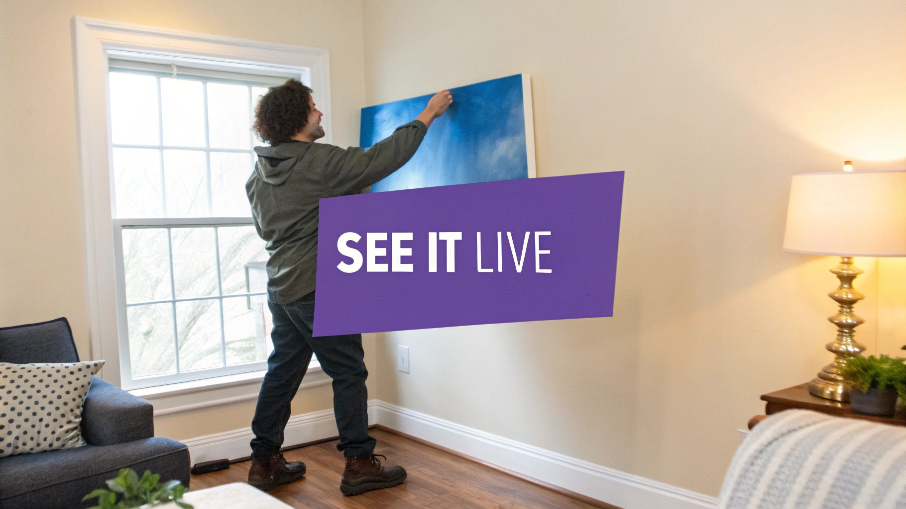 A man hangs a blue painting on a beige wall, with a 'SEE IT LIVE' banner.