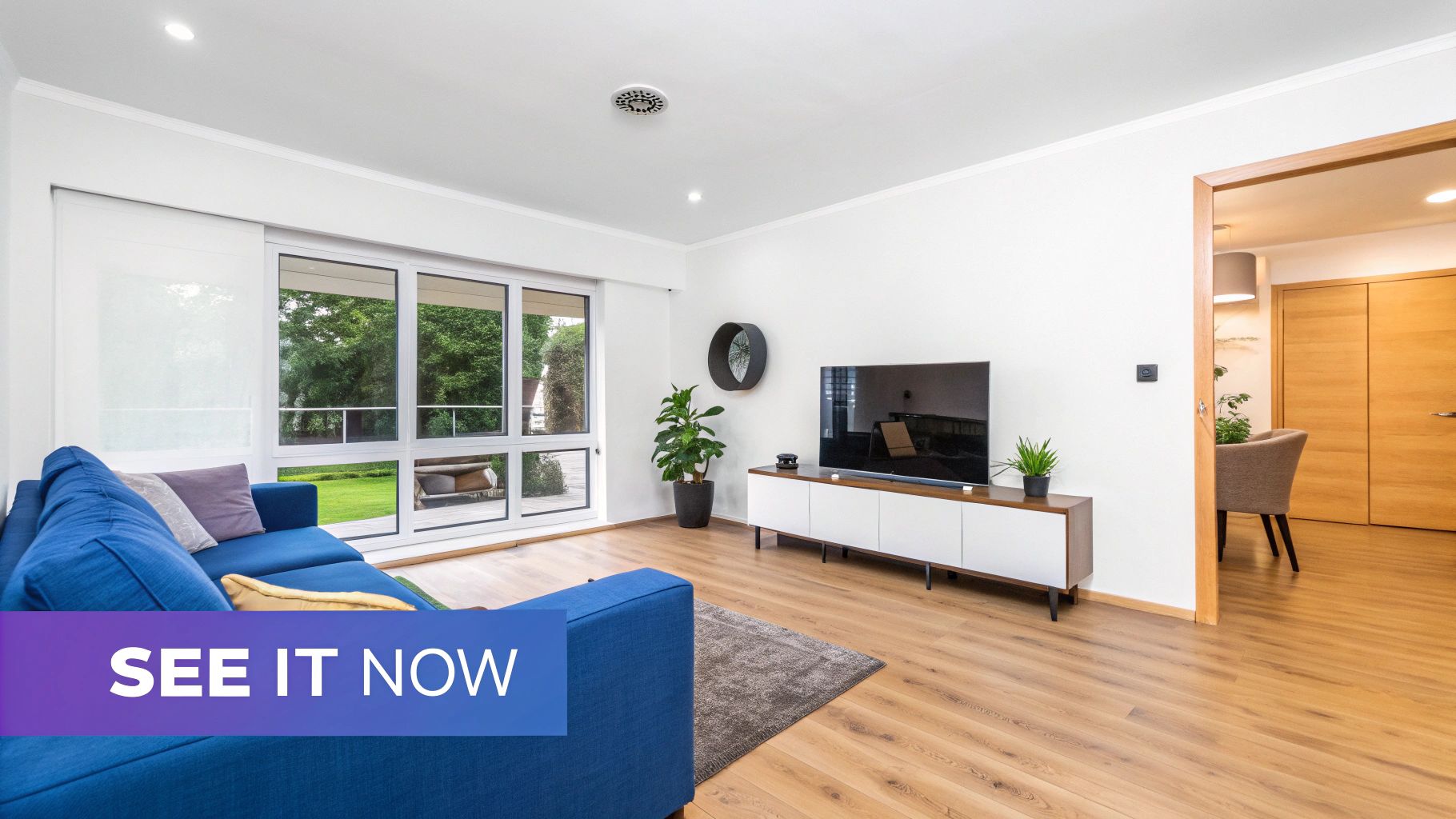 A bright, modern living room featuring a blue sectional sofa, large windows, a TV, and potted plants.