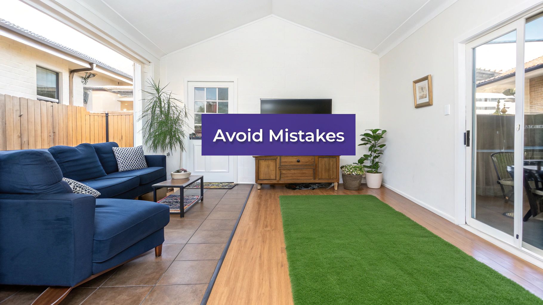 A bright sunroom with blue sectional sofa, a wooden TV console, green rug, and mixed flooring.