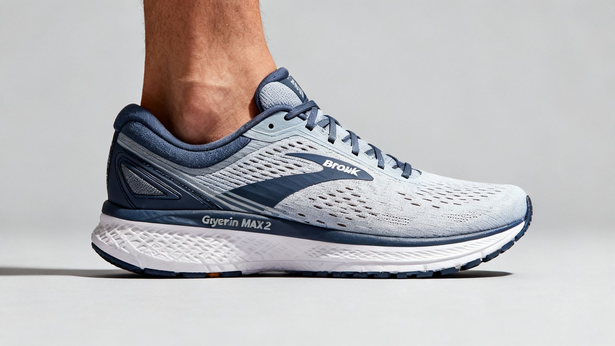 Close-up of a foot wearing a light gray and navy sneaker with "Browk Glycerin MAX 2" text. The background is plain gray.