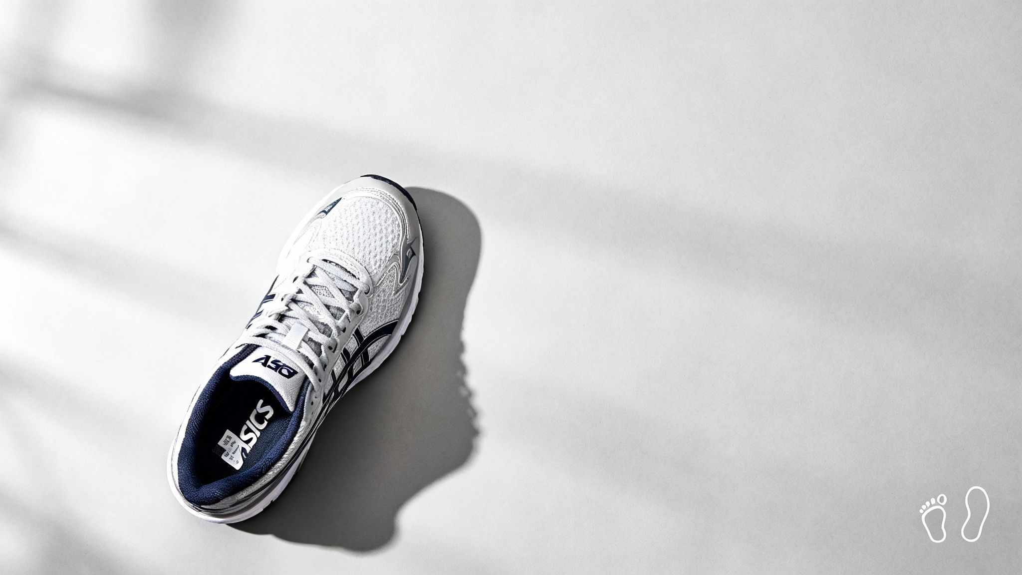 White Asics running shoe on a light gray background with soft shadows. The shoe displays the brand logo and blue details. Minimalist vibe.