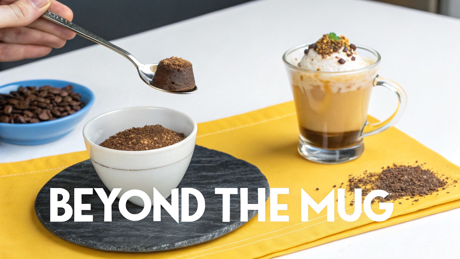 A hand holds a spoon with a small chocolate cake over a bowl of coffee grounds, next to a layered coffee drink with cream.