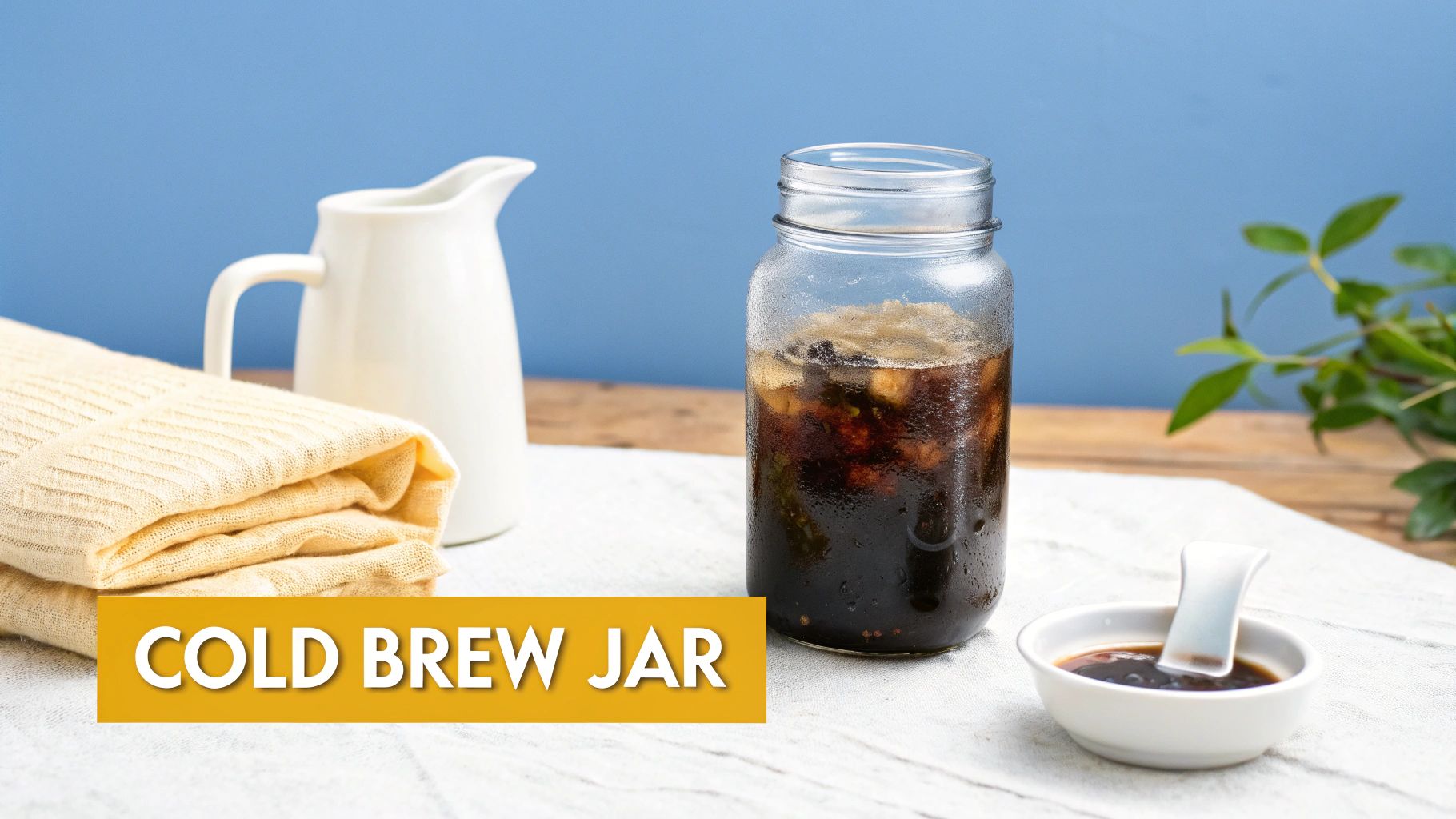 A glass jar of cold brew coffee with ice, alongside a white pitcher and a folded towel on a table.