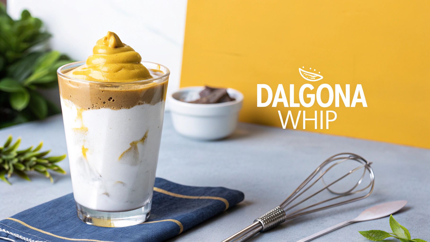 A refreshing Dalgona coffee with golden whipped topping, a whisk, and green leaves.