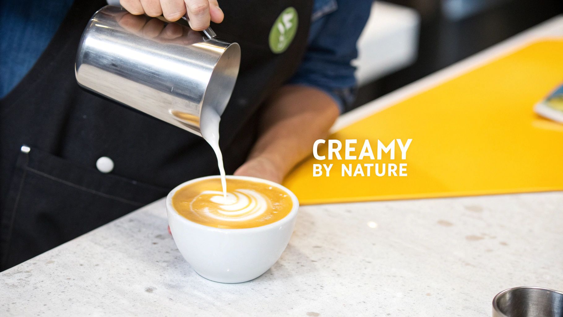 A barista expertly pours frothed milk from a silver pitcher into a white coffee cup, crafting beautiful latte art.