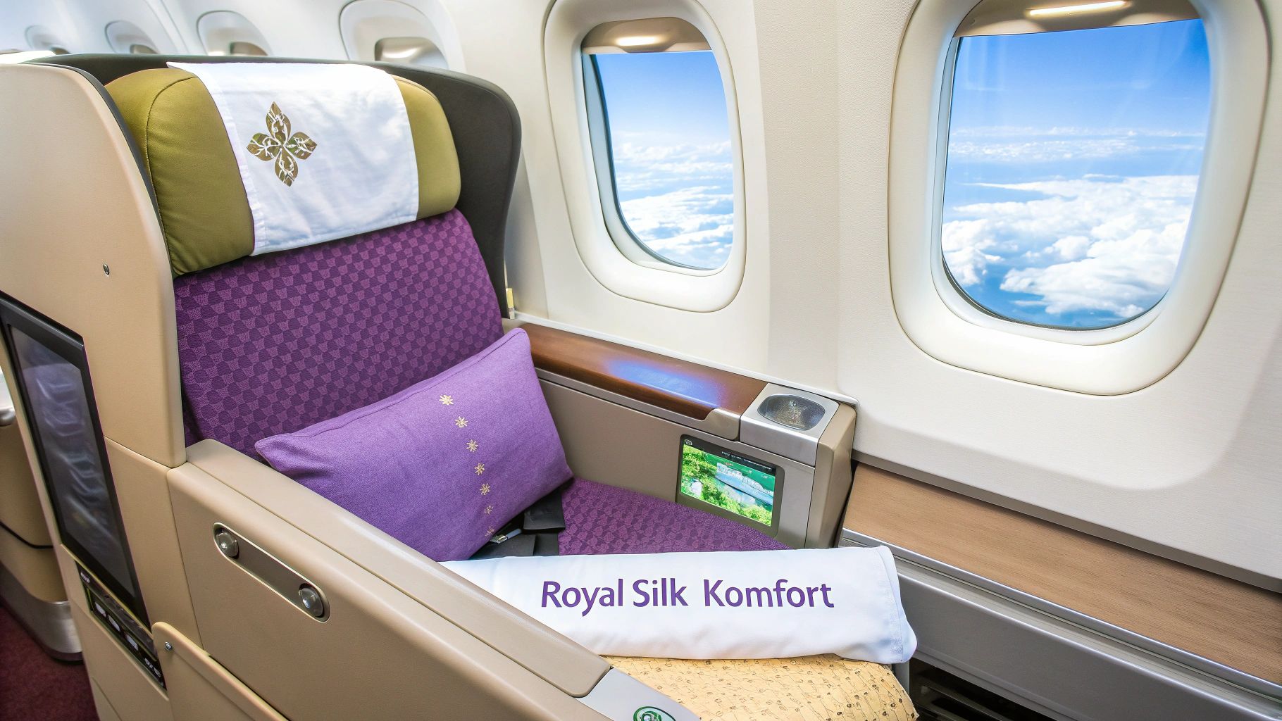 Ein Business-Class-Sitz in einem Flugzeug mit lila Kissen und Blick aus dem Fenster auf den blauen Himmel und Wolken.