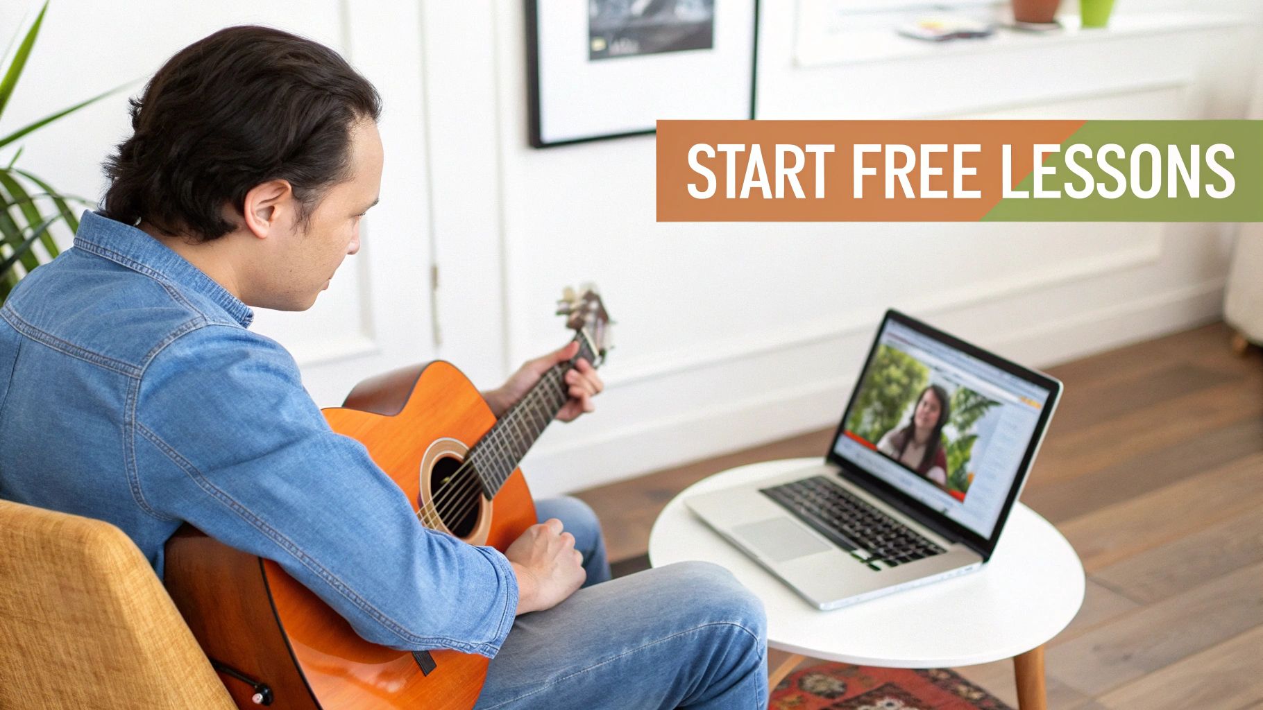 A man in a denim shirt practices acoustic guitar while watching online lessons on a laptop.