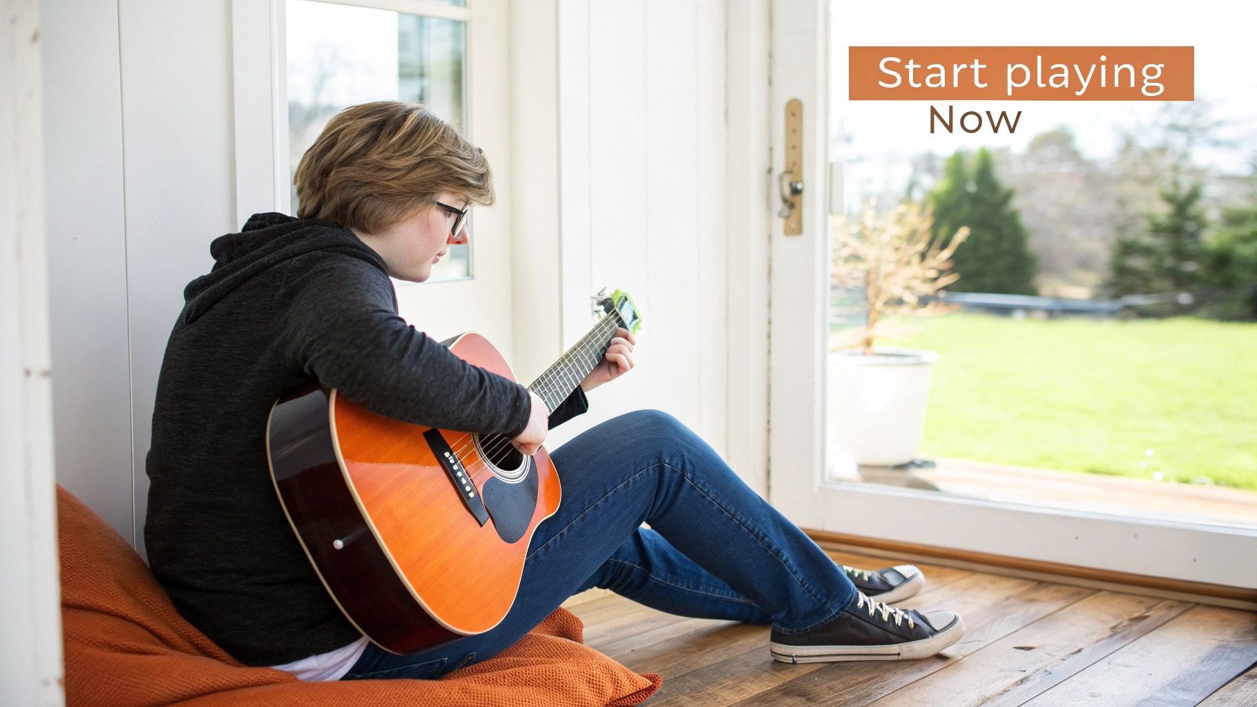 A young person sits on a cushion by a sunlit window, focused on playing an acoustic guitar.