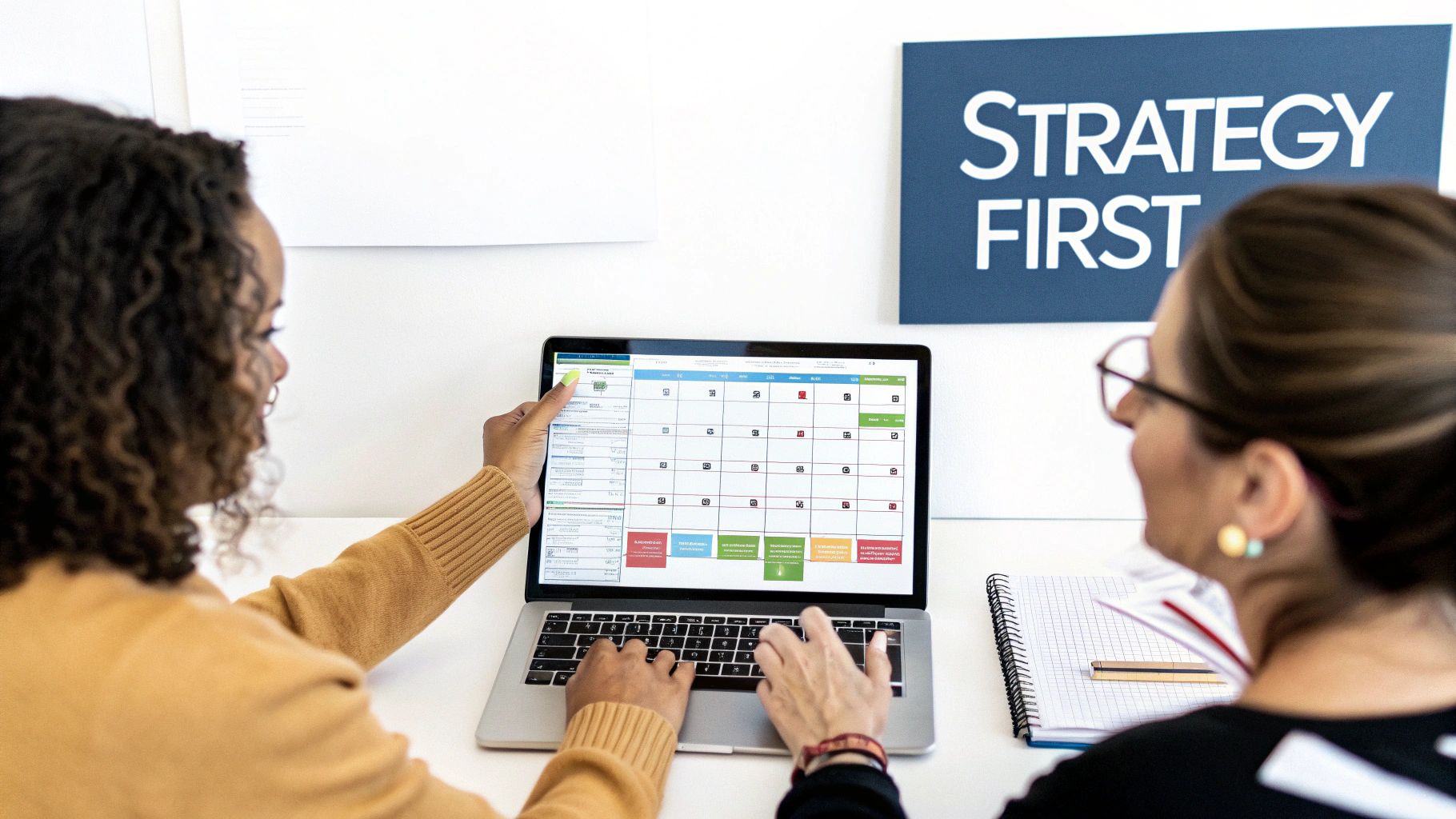 Two women collaborate on a laptop displaying a content calendar, with a 'Strategy First' sign in the background.