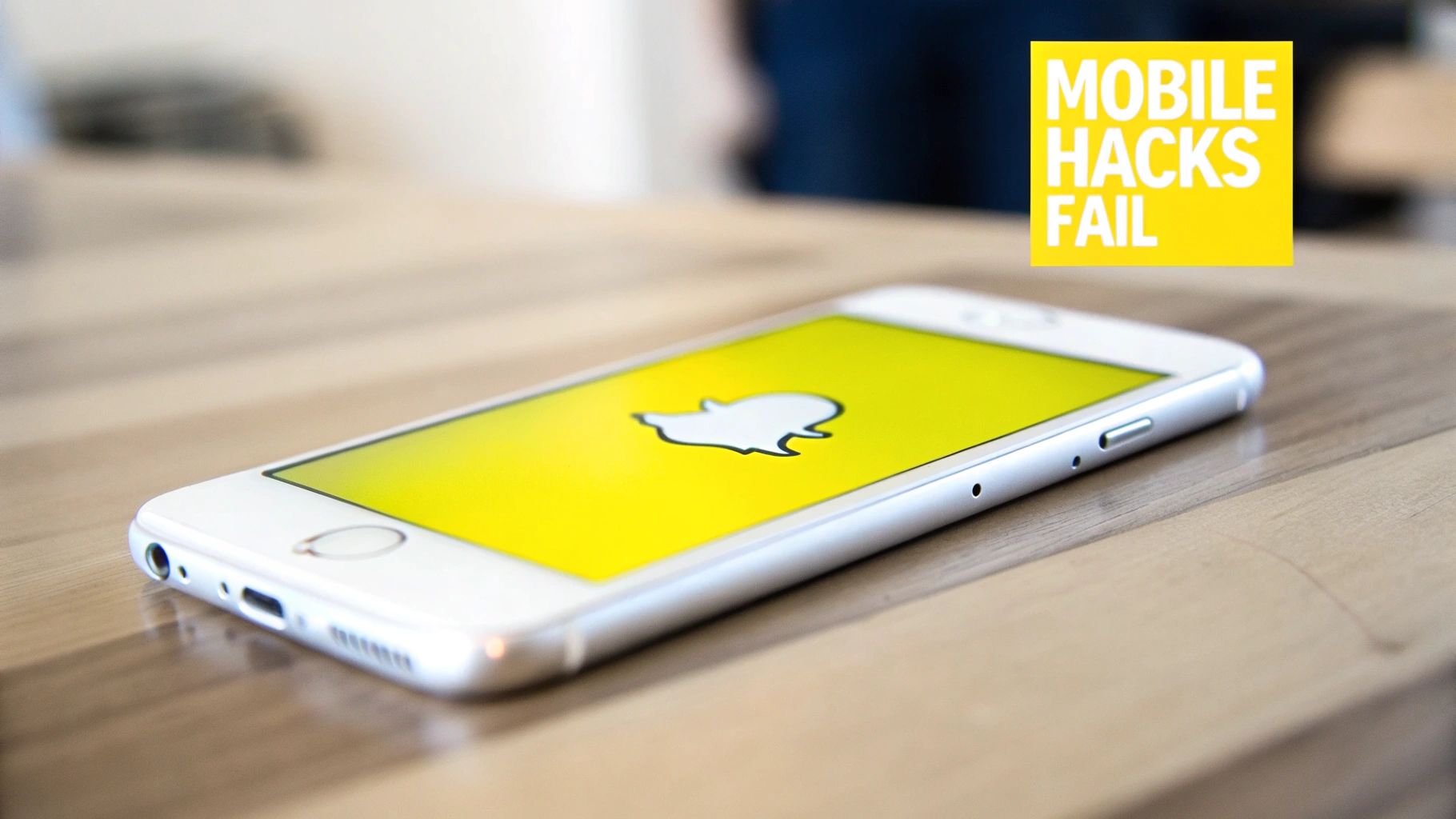 A white iPhone with the Snapchat ghost logo on its screen, lying on a wooden table, with a 'Mobile Hacks Fail' caption.