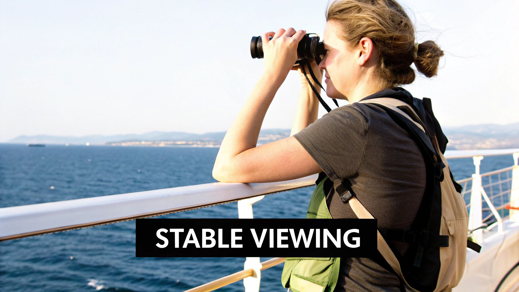 A woman on a cruise ship deck uses binoculars to view the ocean and distant coastline.