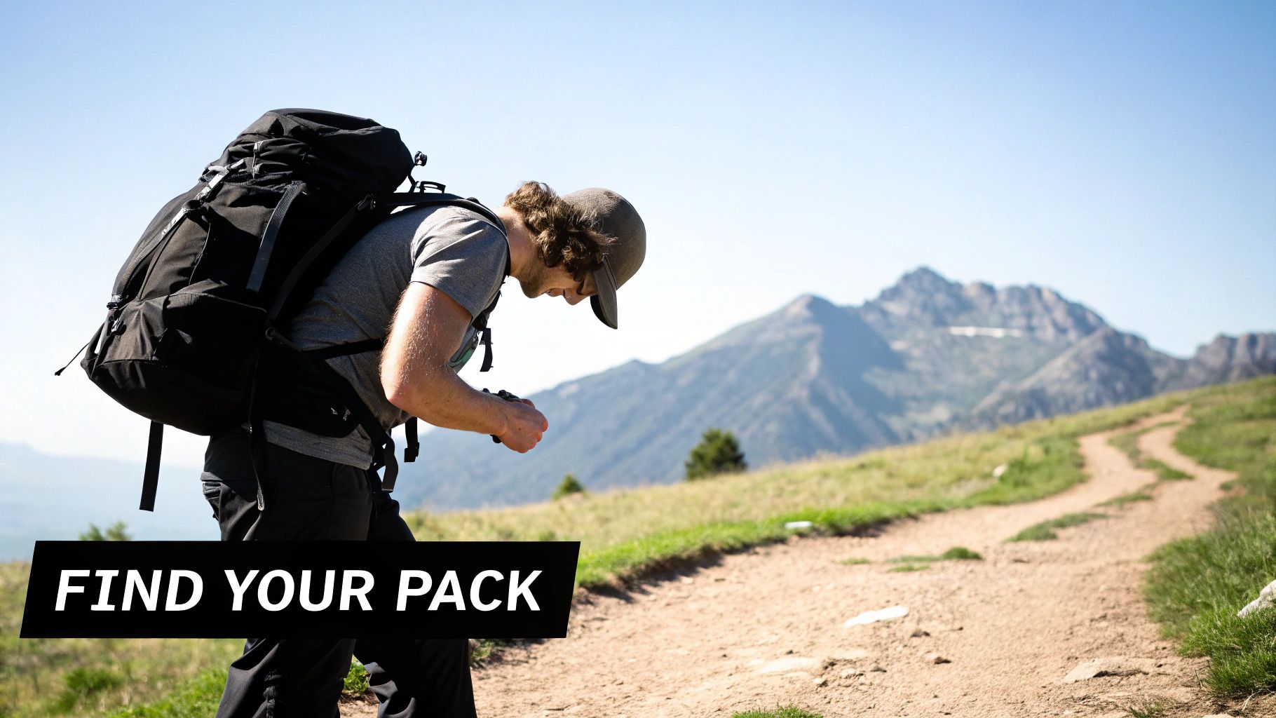 A man with a large black trekking backpack walks on a dirt trail in a sunny mountain landscape.