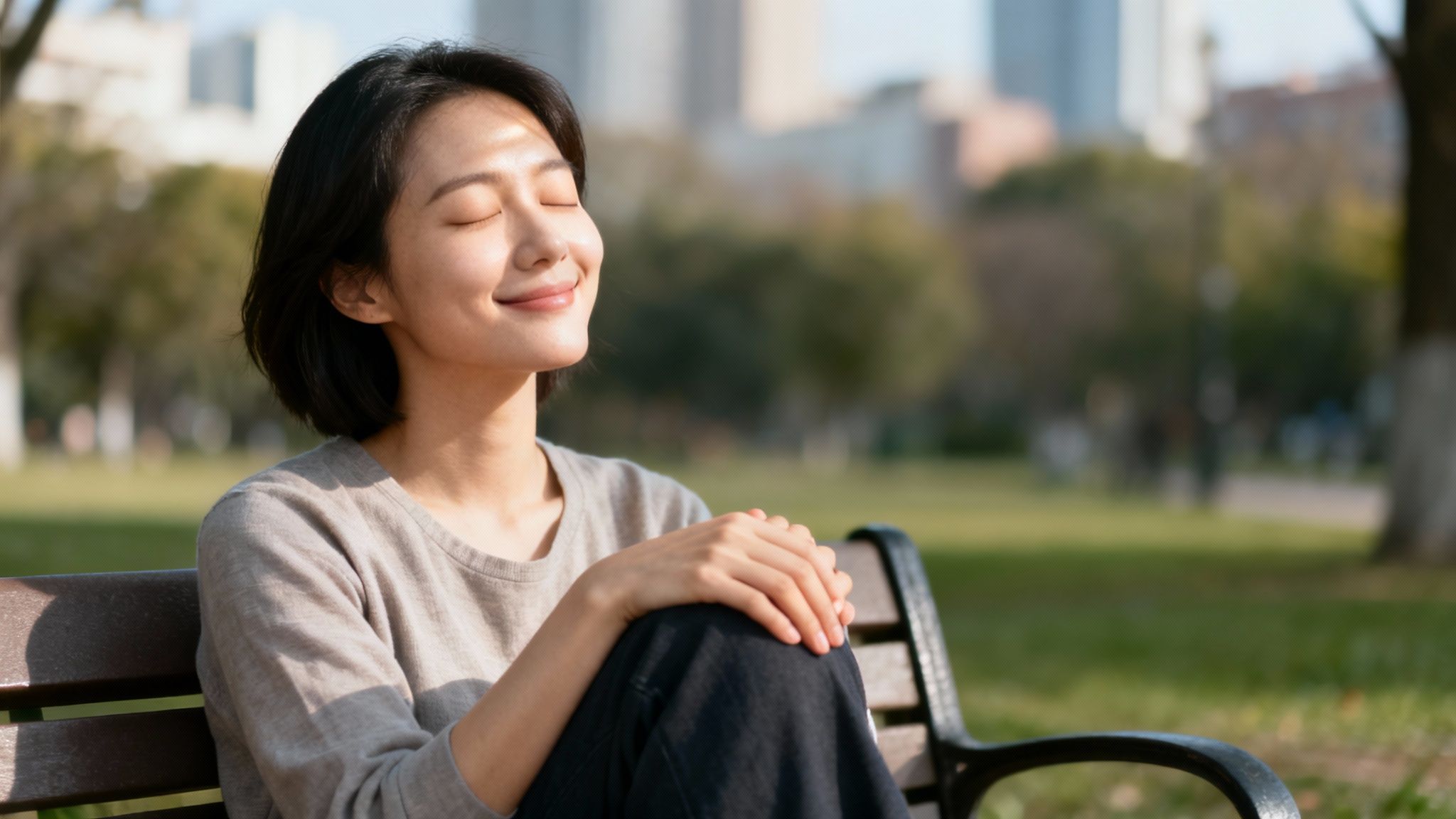 A young Asian woman enjoying sunlight on a park bench, eyes closed, smiling peacefully.