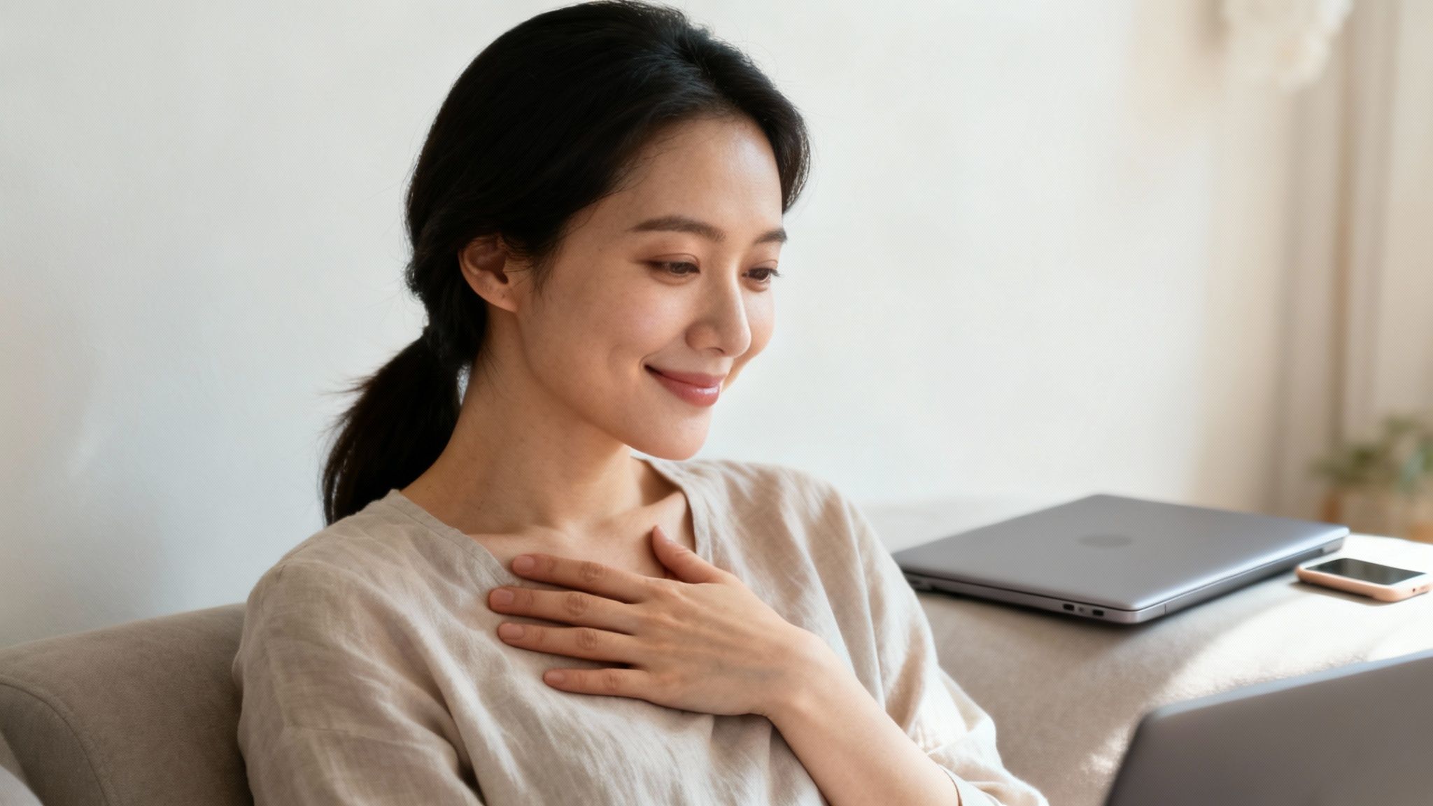 Smiling Asian woman watches a guided meditation on her laptop in a peaceful home.