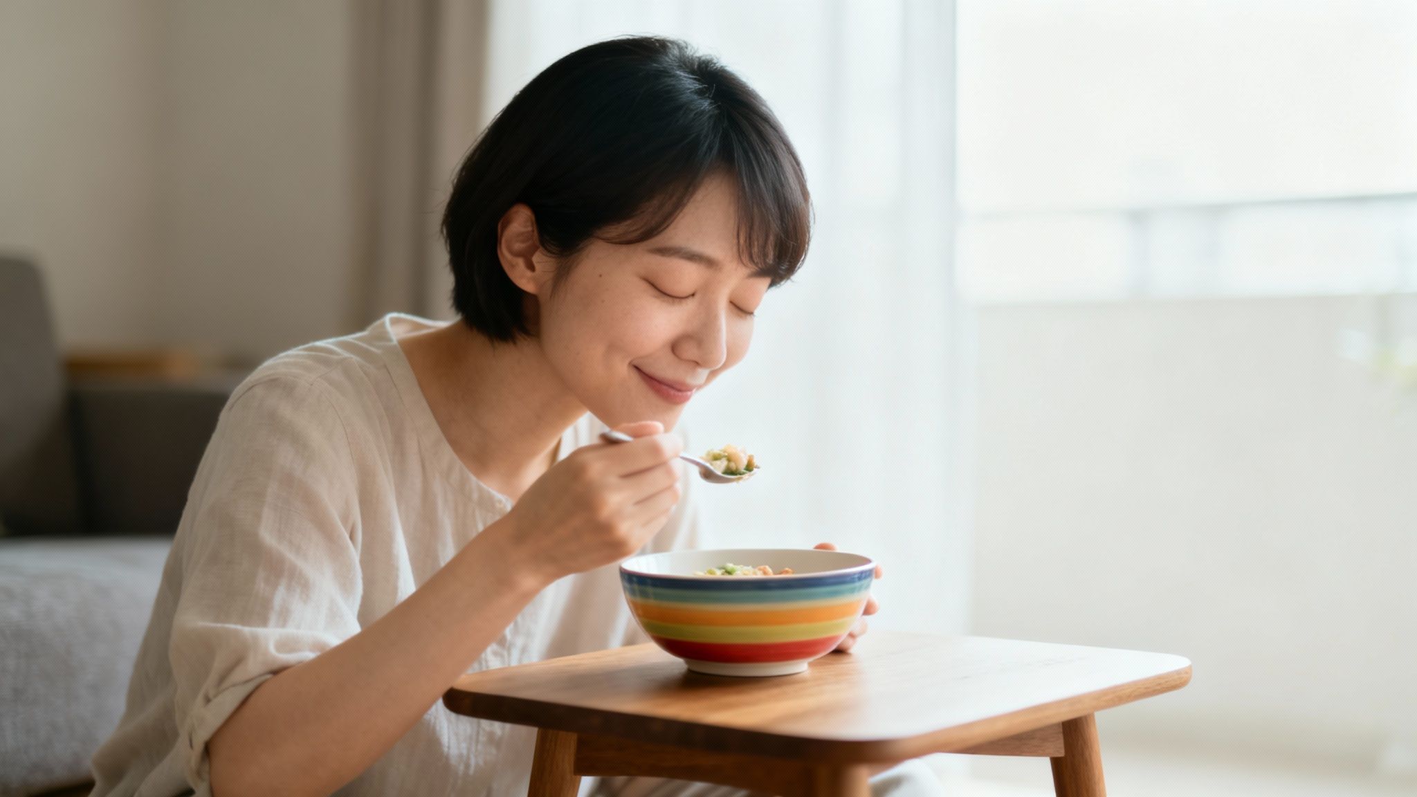 A young Asian woman with her eyes closed, savoring a spoonful of food from a colorful bowl.