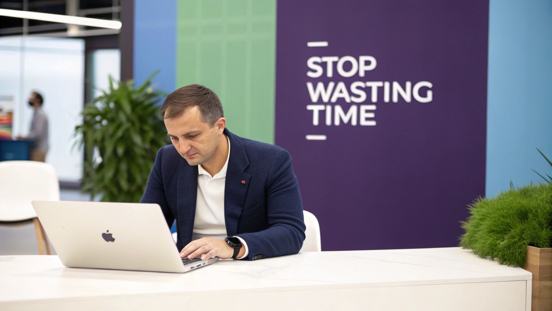 A focused man in a blue blazer works on a MacBook laptop at a modern office desk with a 'STOP WASTING TIME' sign.