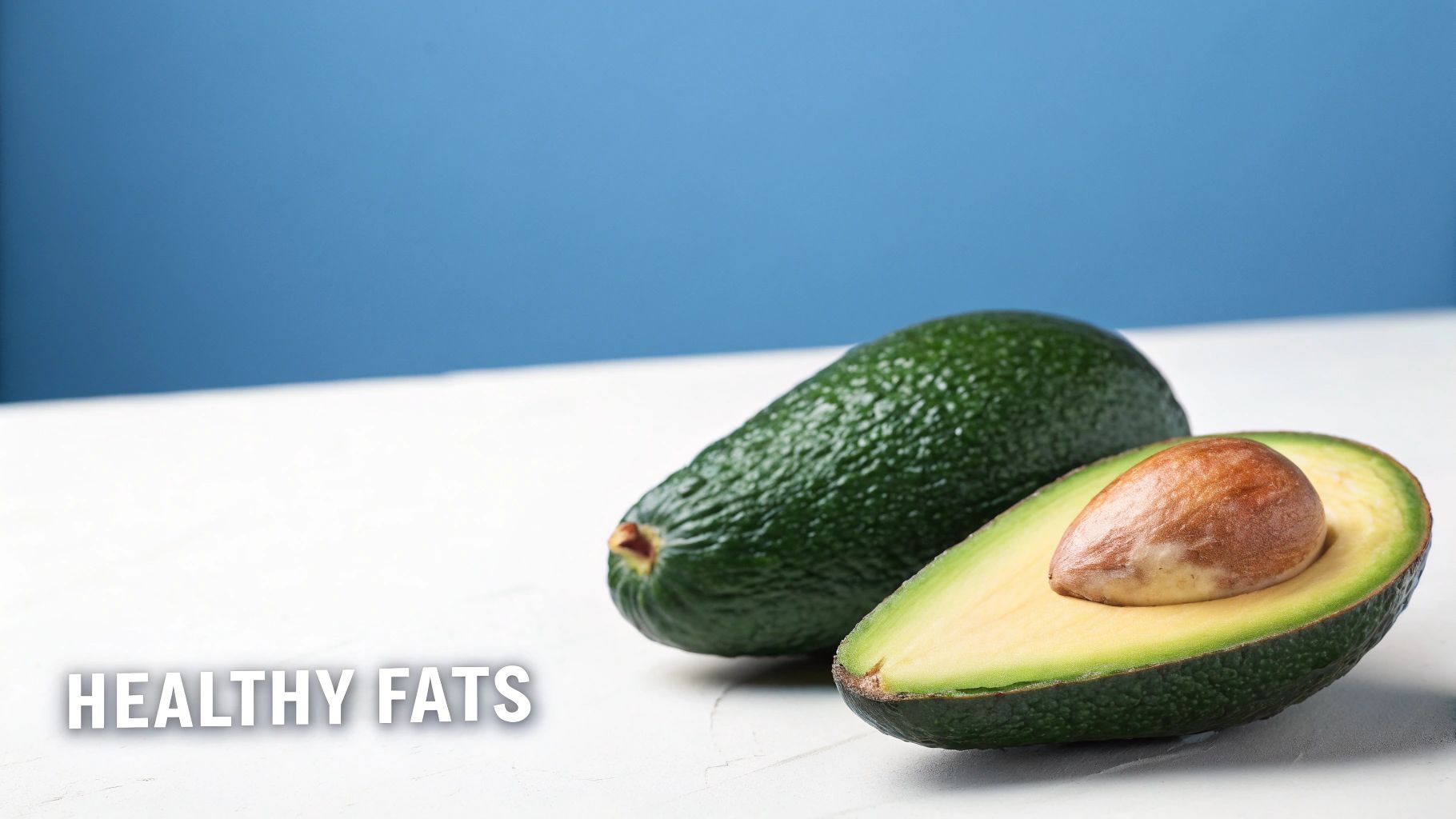 A whole and a halved avocado with its pit on a white surface, against a blue background, promoting healthy fats.