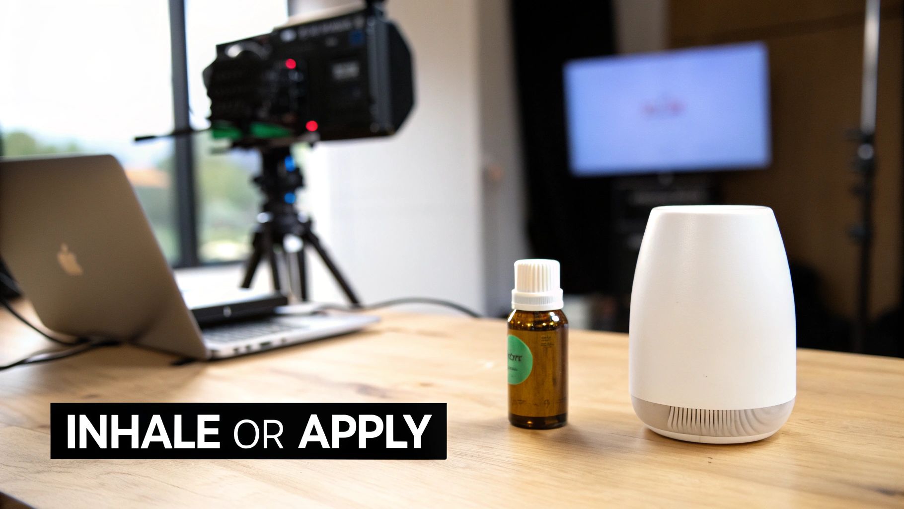 An essential oil bottle and diffuser on a wooden table in a room with a camera and laptop.