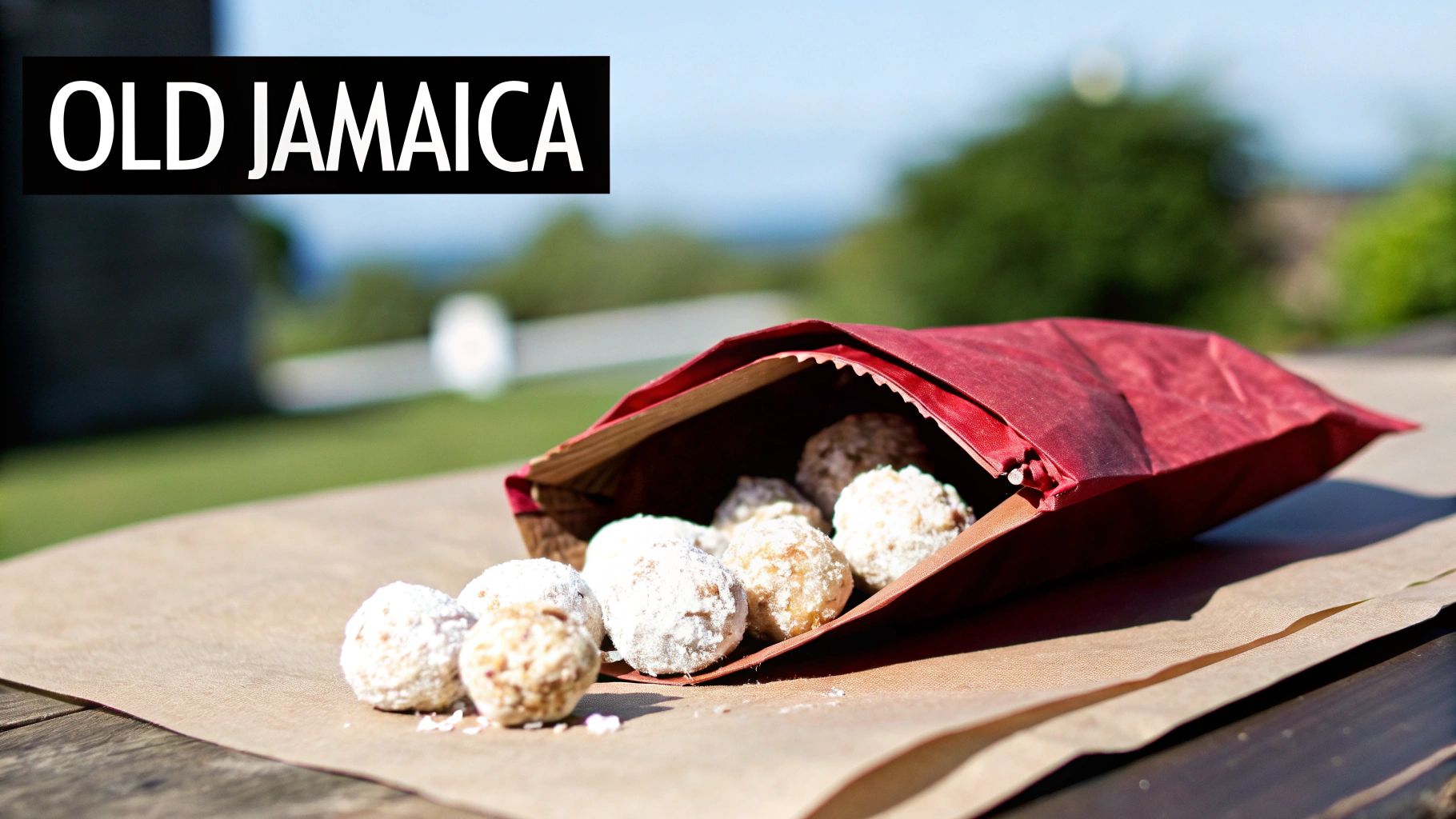 Sugar-dusted round sweets spilling from a red paper bag onto a brown paper surface outdoors under the text 'OLD JAMAICA'.