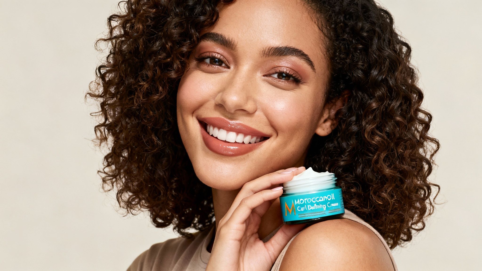Una mujer feliz con cabello rizado sonríe mientras sostiene un frasco de Crema Definidora de Rizos Moroccanoil.