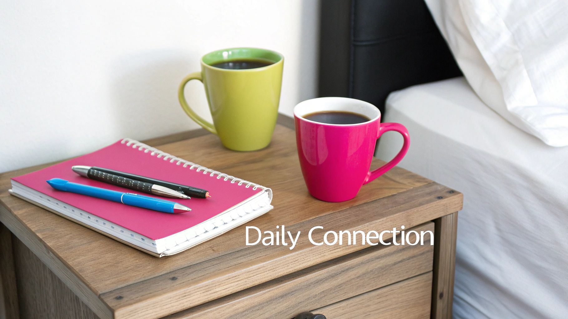 Two colorful coffee mugs and a pink notebook with pens on a wooden nightstand next to a bed.