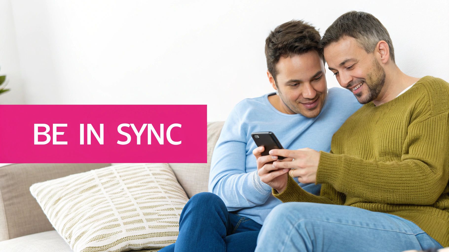 Two smiling men on a couch looking at a smartphone together, with 'BE IN SYNC' text.