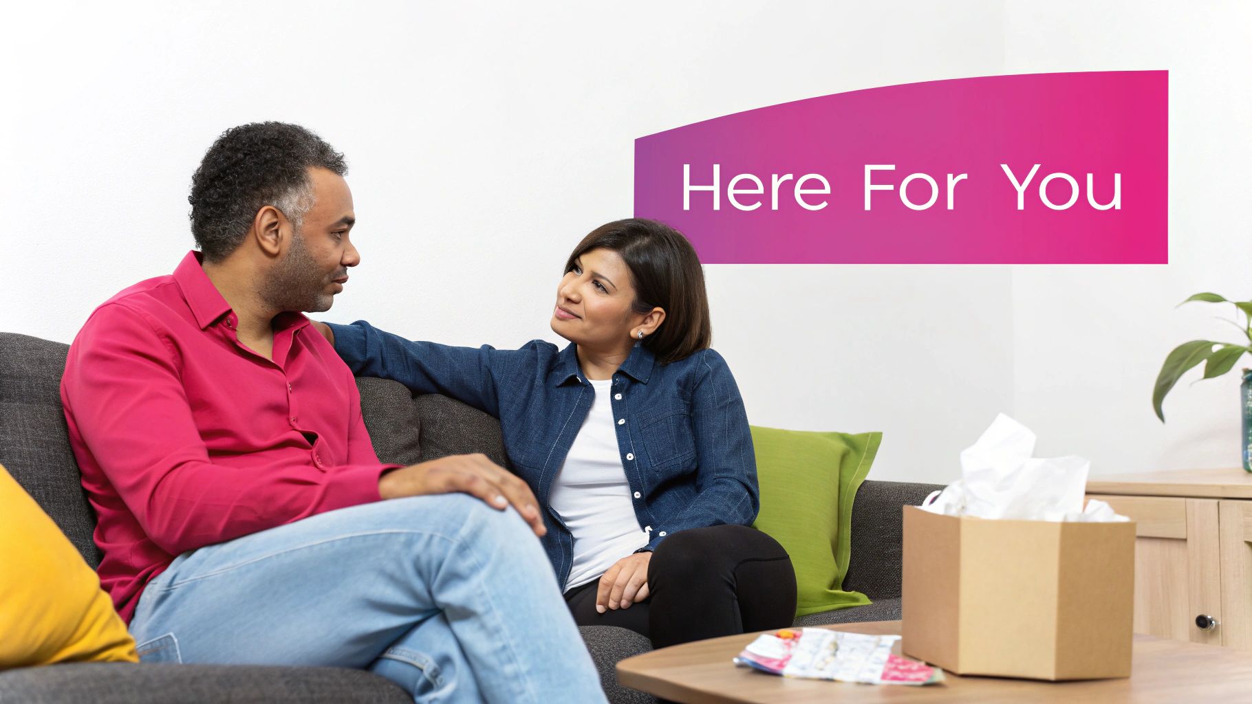A woman comforts a man on a couch, with a tissue box nearby and 'Here For You' banner.