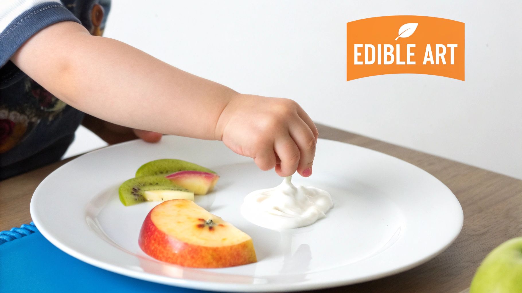 A toddler's hand dips apple and kiwi slices into yogurt on a white plate.