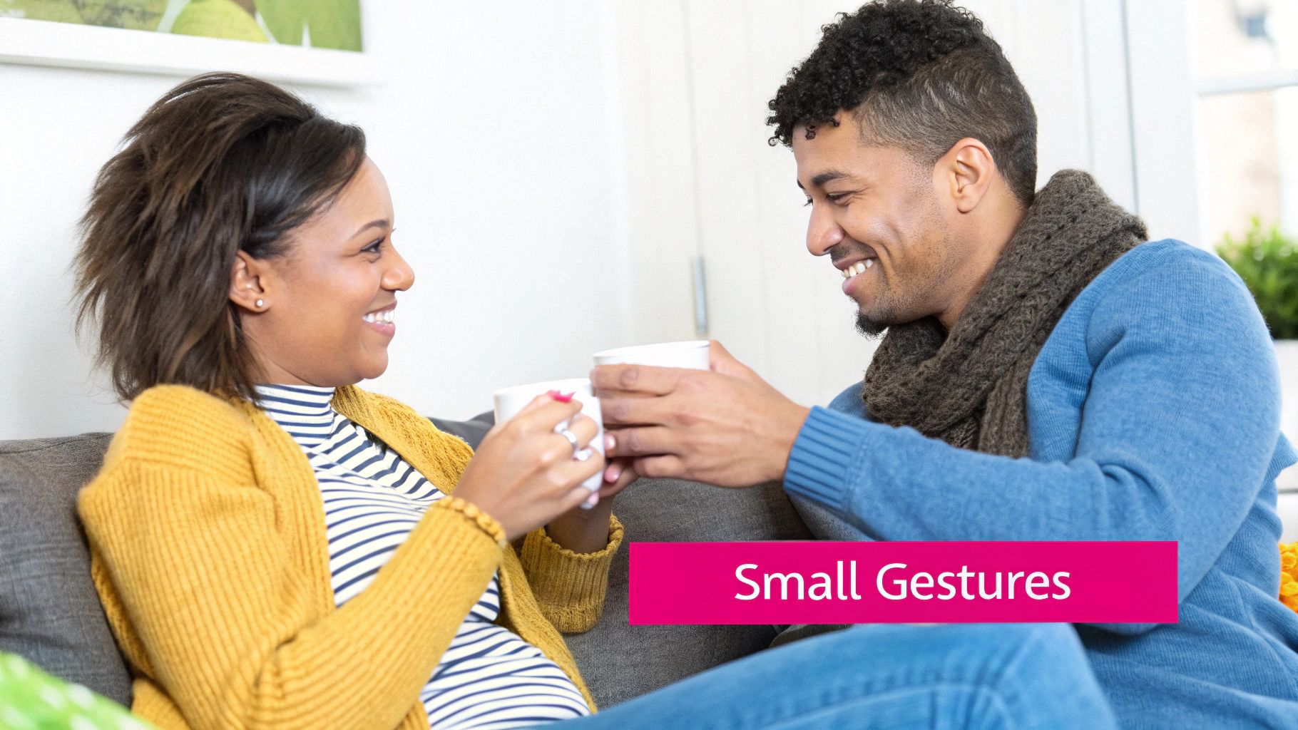 A happy couple sitting on a sofa, smiling at each other and holding white mugs.