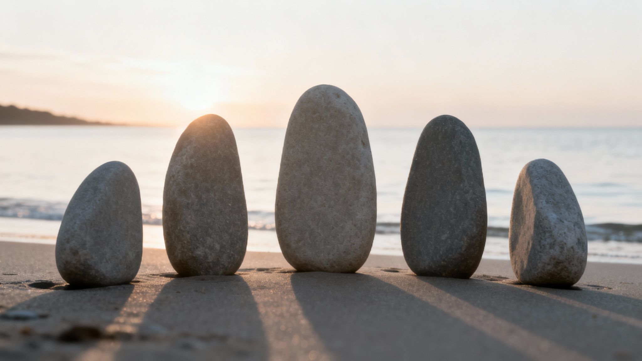 Five balanced grey stones stand upright on a sandy beach at sunset, casting long shadows.