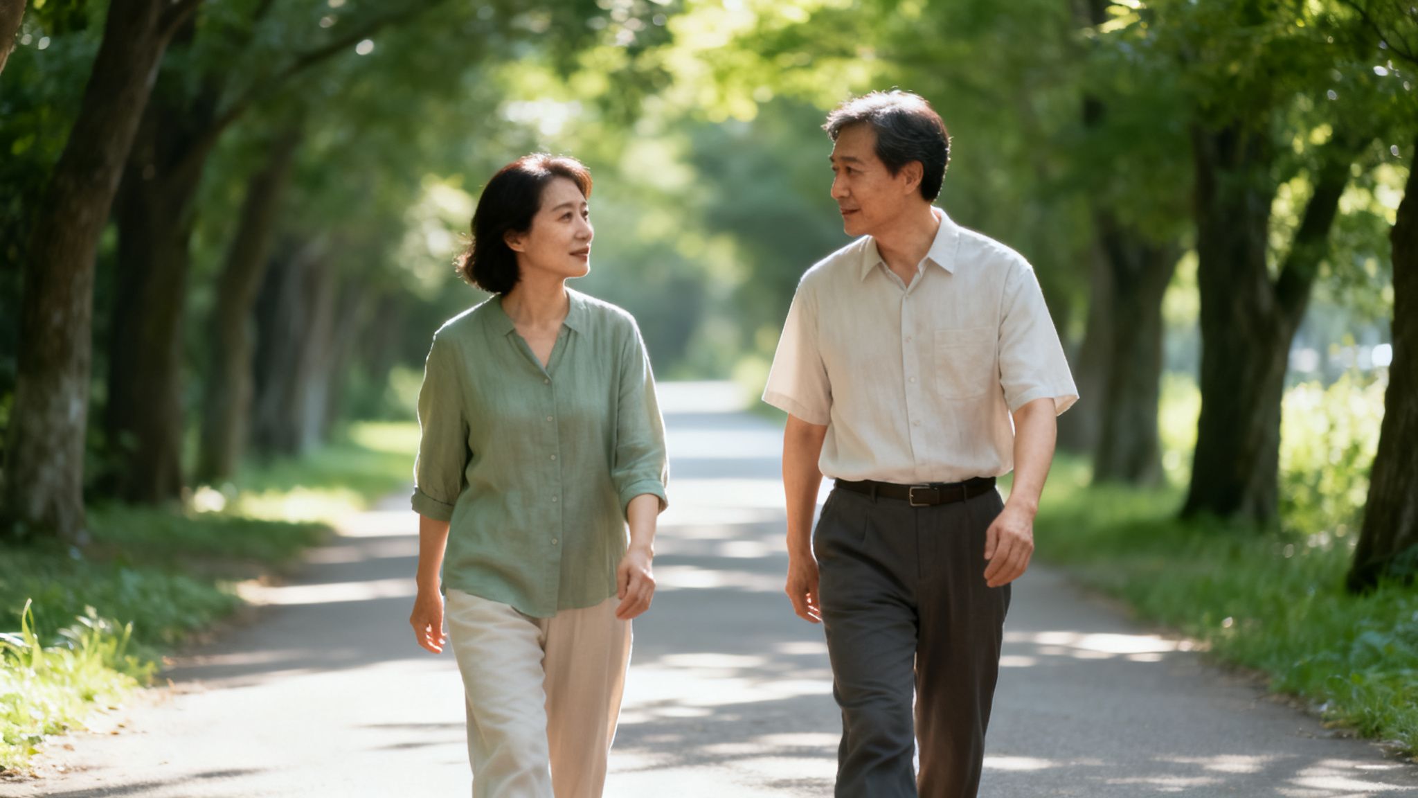 A mature Asian couple walks and converses on a sunny, tree-lined path.
