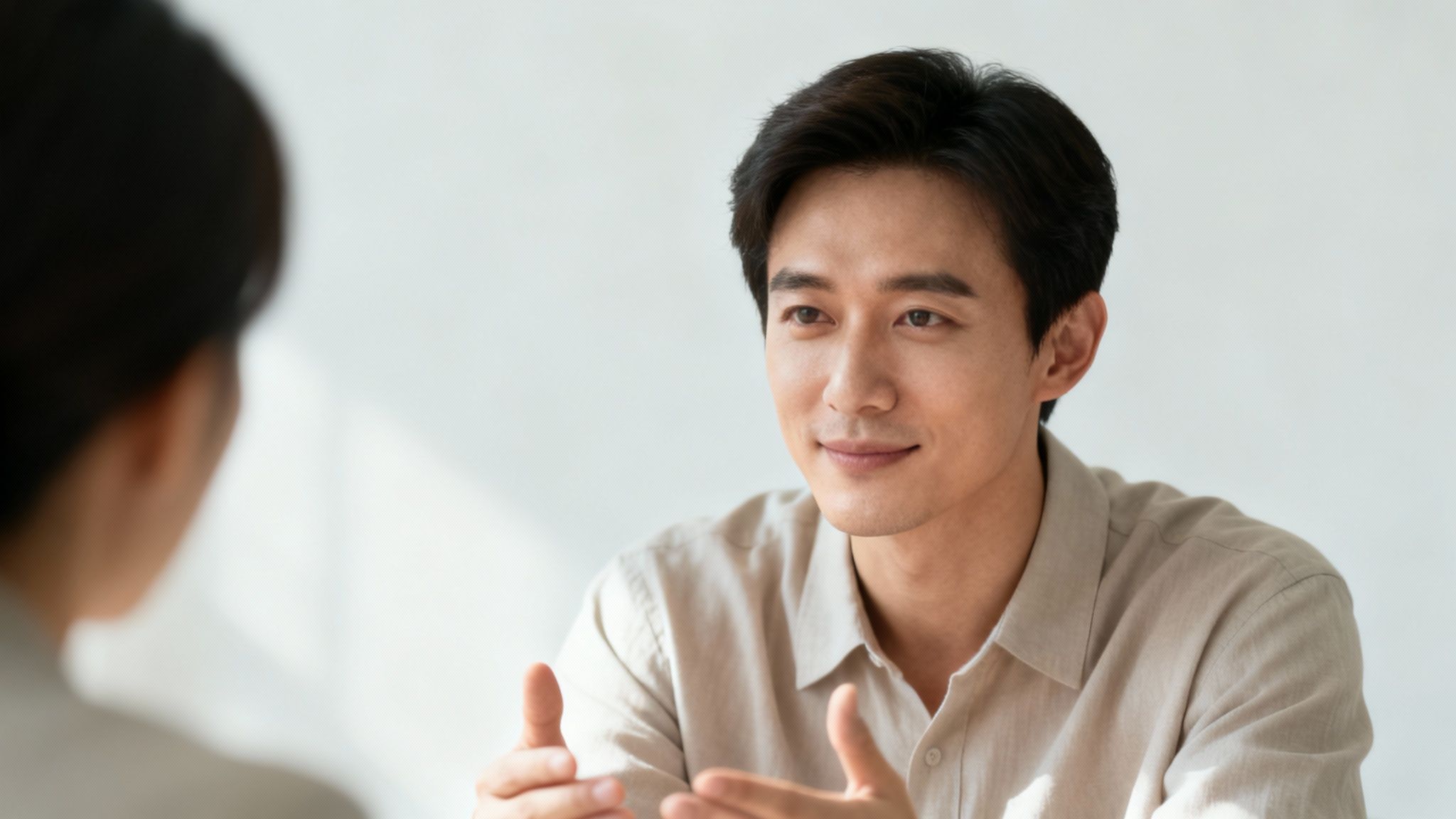 A smiling Asian man in a beige shirt talks and gestures to a person across from him.