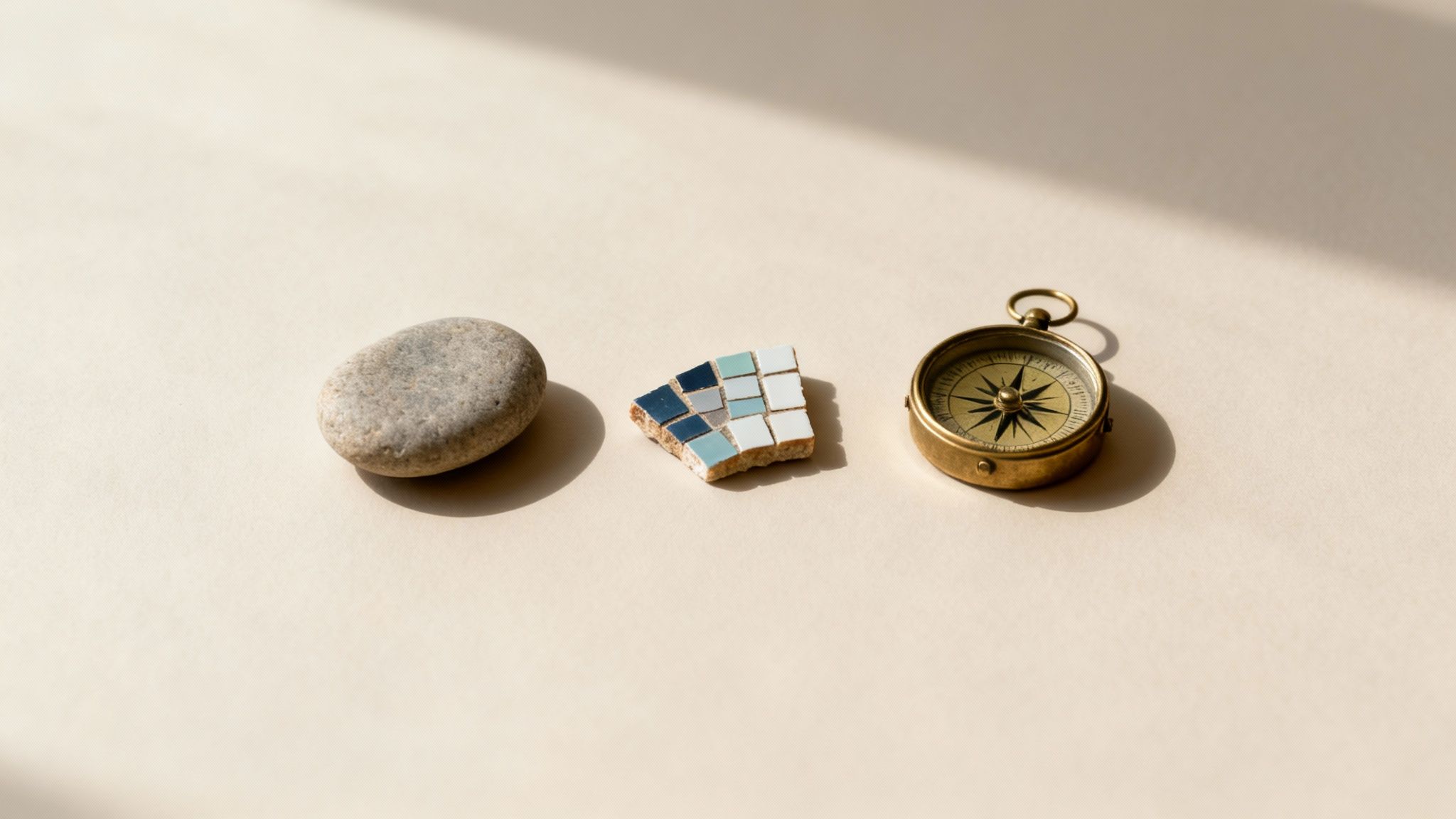A smooth grey stone, a blue mosaic tile shard, and a brass compass on a beige surface.