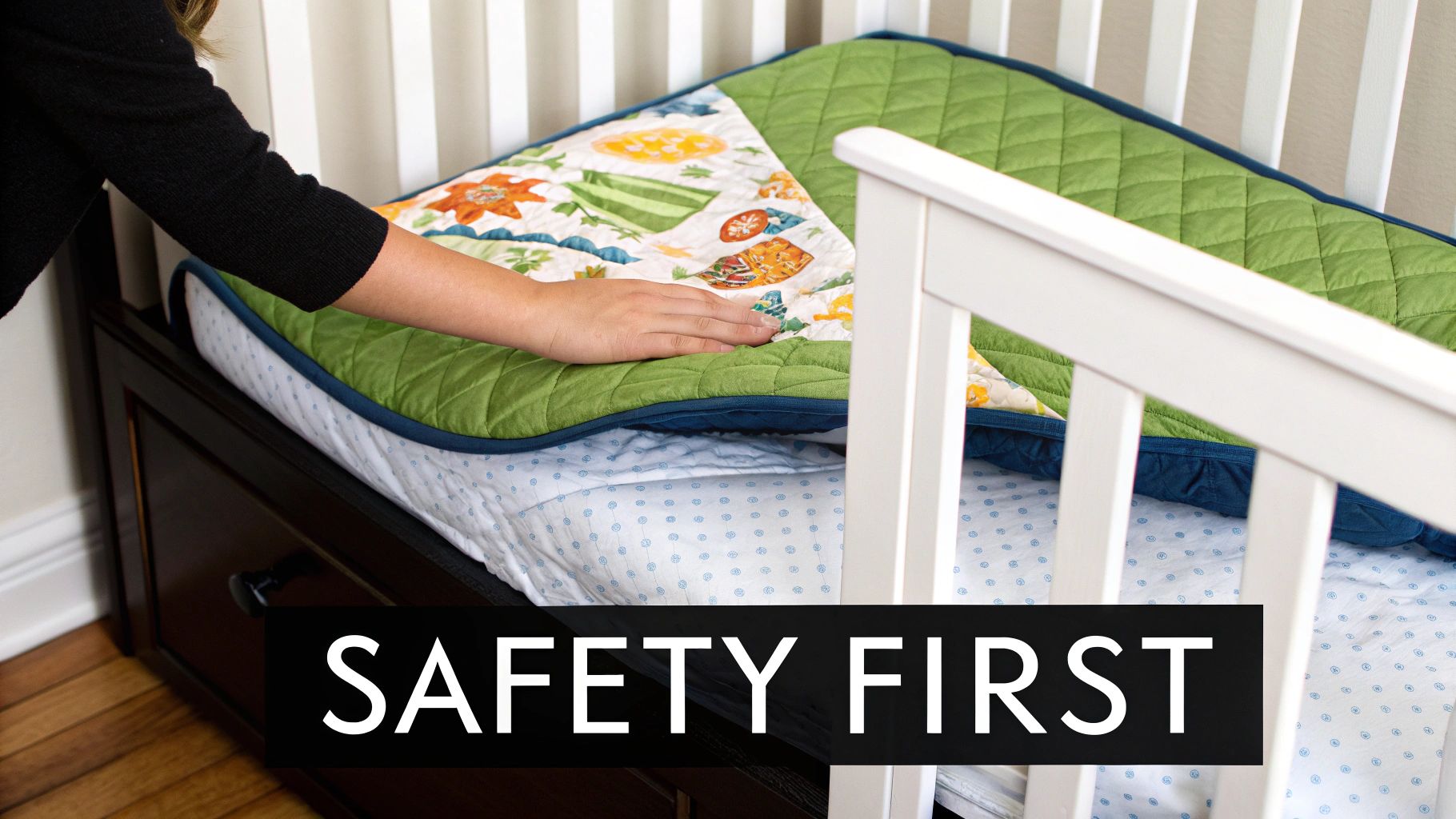 A person's hand adjusts a green and colorful quilt inside a white baby crib with a dotted sheet.