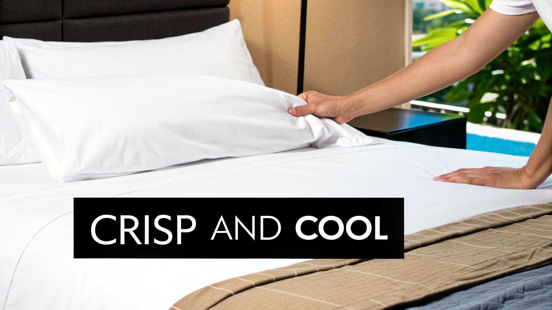 A hand adjusting a white pillow on a freshly made bed with crisp sheets in a hotel room.