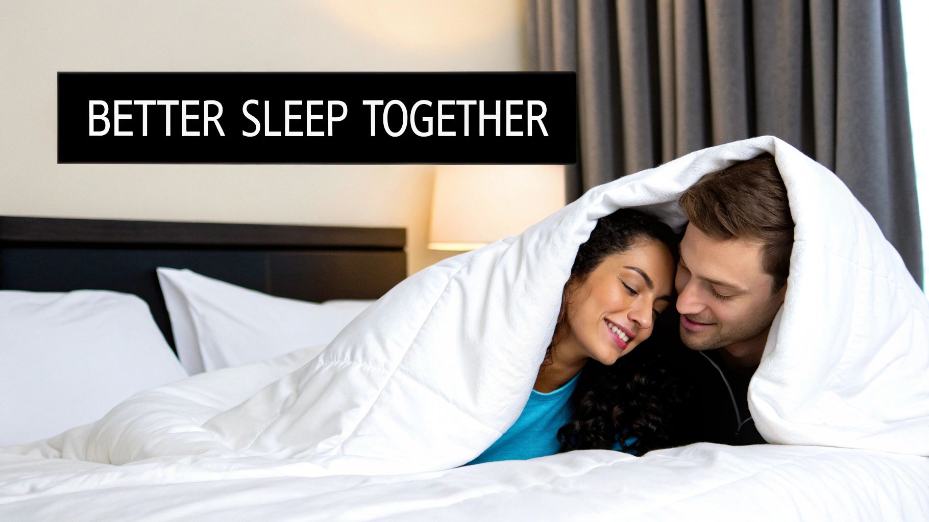 A happy couple snuggled under a white duvet in bed, smiling at each other.