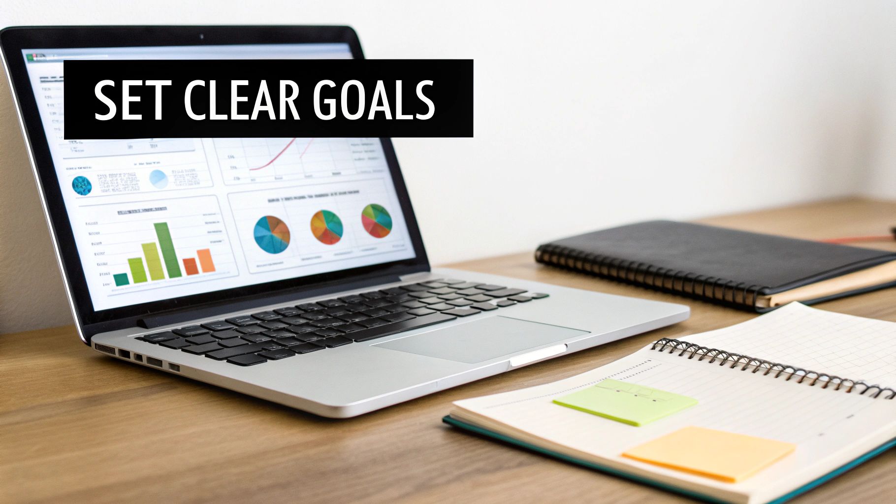 A laptop showing charts and graphs, with a notebook and sticky notes on a desk. Text: SET CLEAR GOALS.