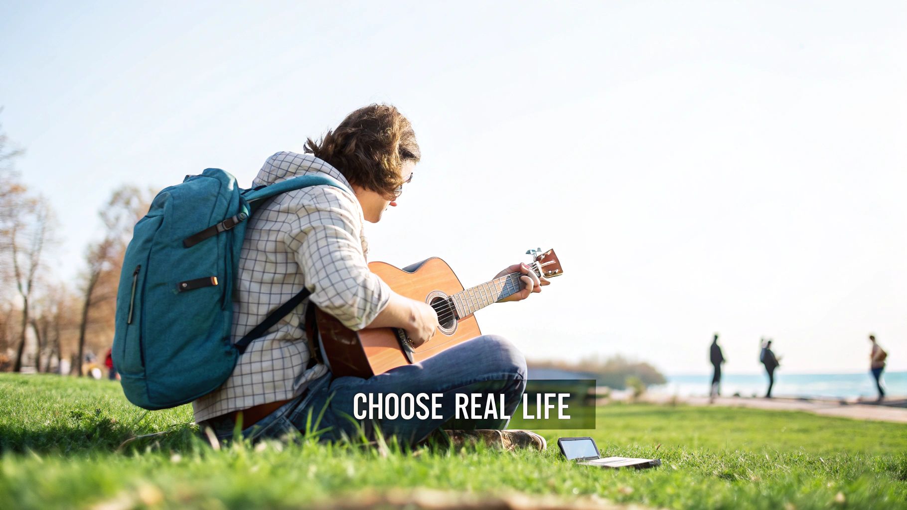 Young person with backpack plays acoustic guitar on grass by the water, near a laptop.