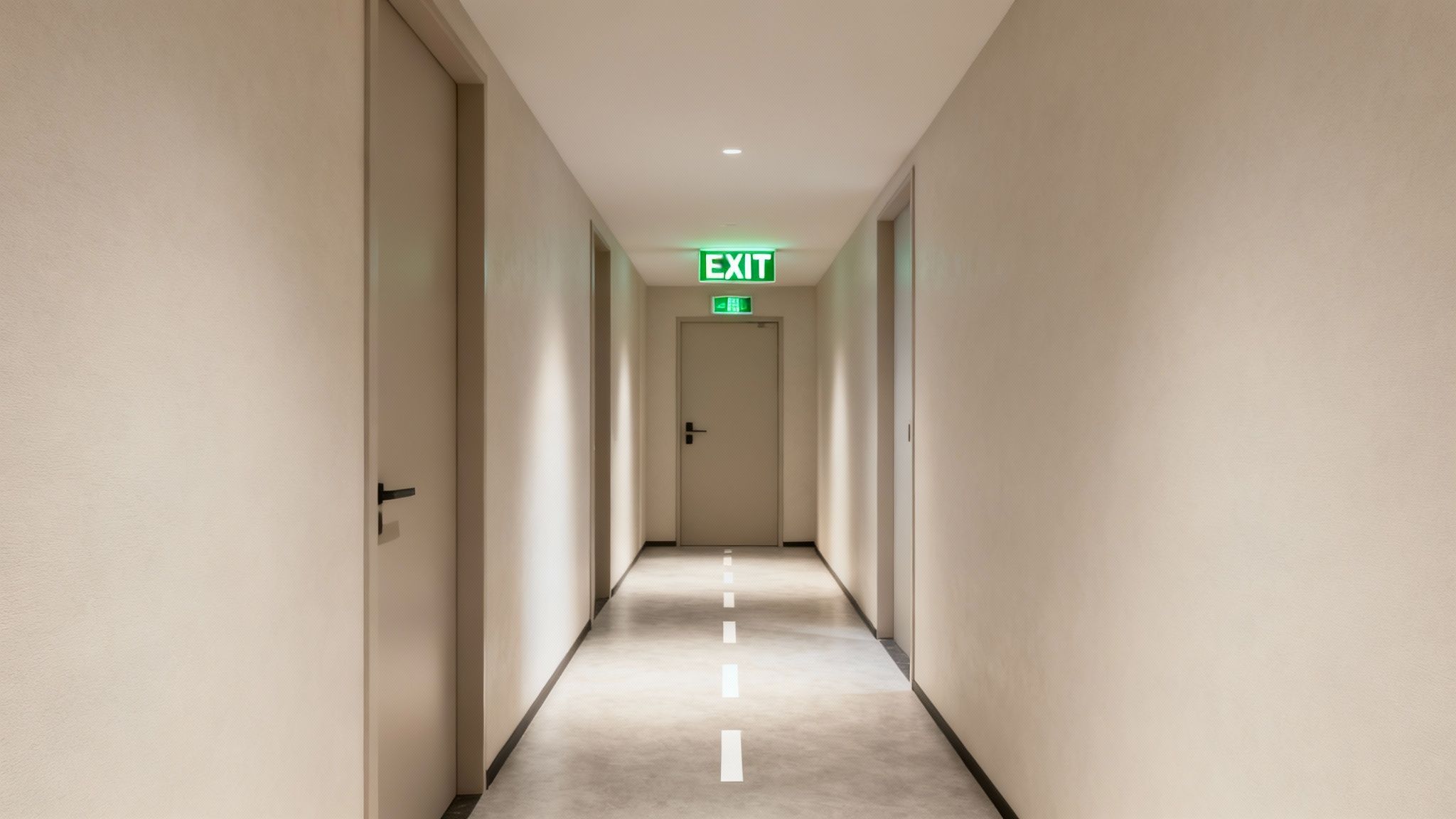 A long, light-colored hallway leads to an emergency exit door with a glowing green sign.