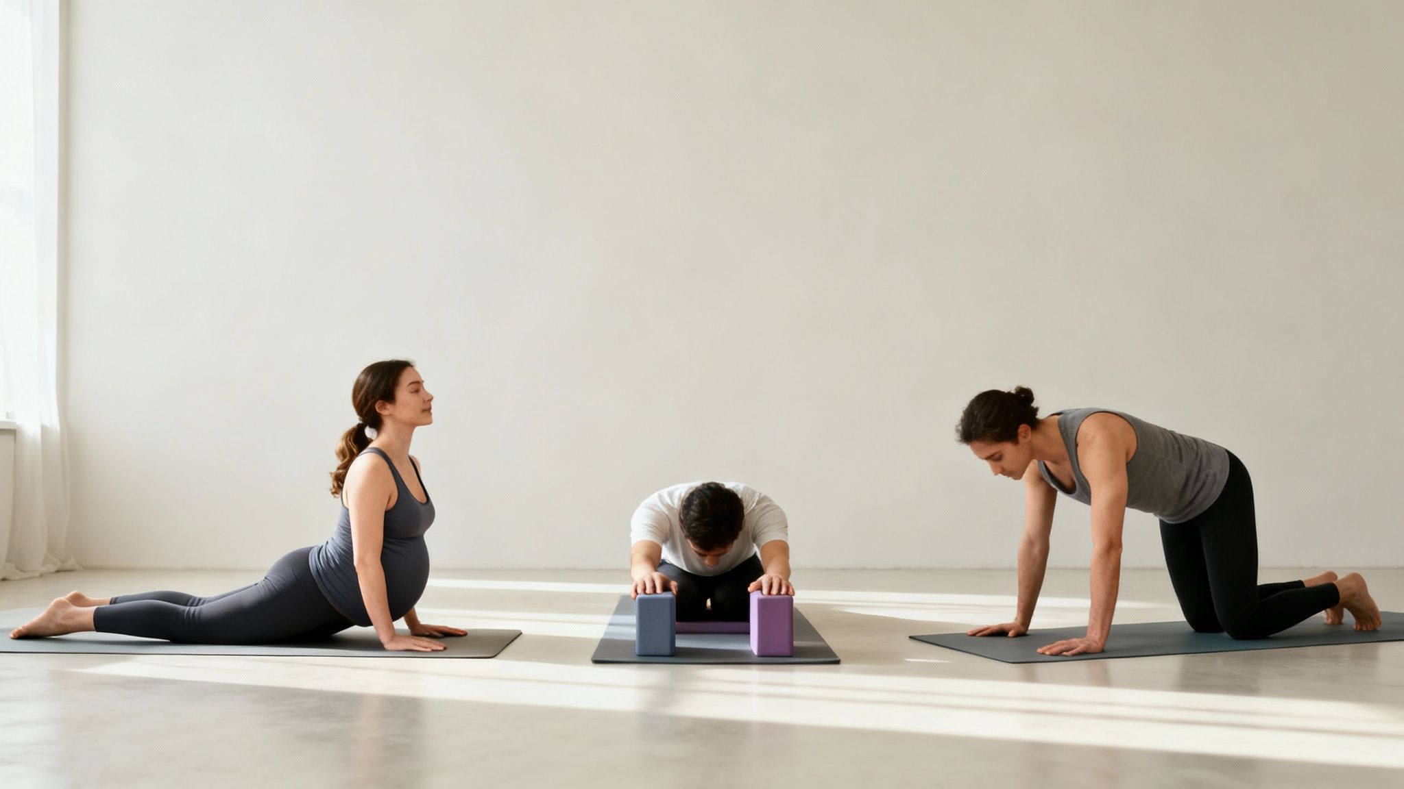 Tři lidé cvičí jógu v jasné místnosti: těhotná žena v kobře, muž s bloky a další na všech čtyřech.