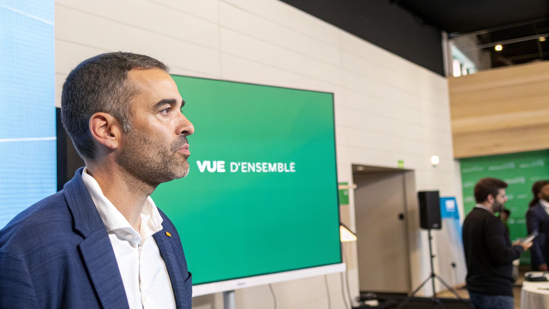 Un homme en veste bleue et chemise blanche, regardant un écran vert affichant "VUE D'ENSEMBLE" lors d'une présentation.