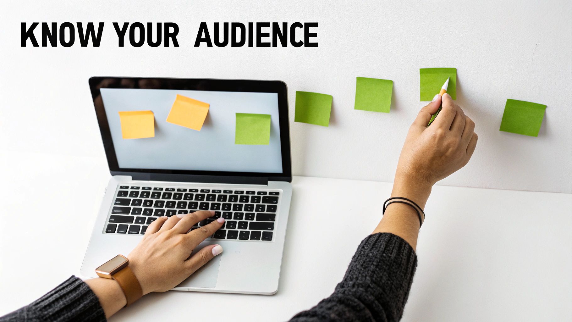 Person typing on laptop while another hand writes on green sticky notes on a white wall, with the text 'KNOW YOUR AUDIENCE'.