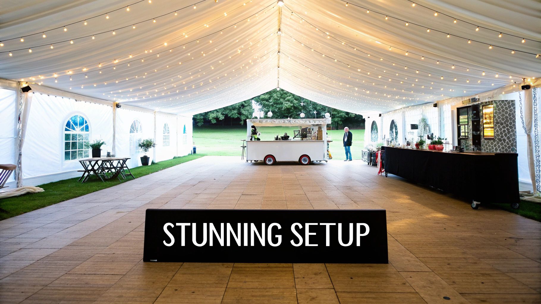 A spacious white marquee tent set up for a wedding or event, featuring string lights, a mobile bar, and a long bar counter.