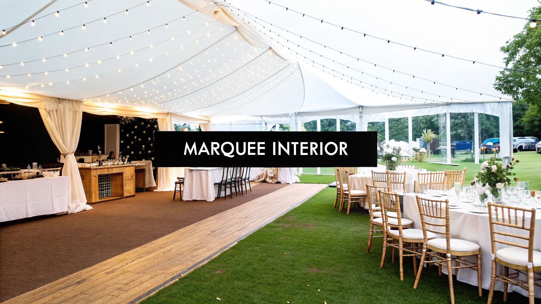 A spacious wedding marquee interior featuring dining tables on grass, a bar area, and elegant string lights.