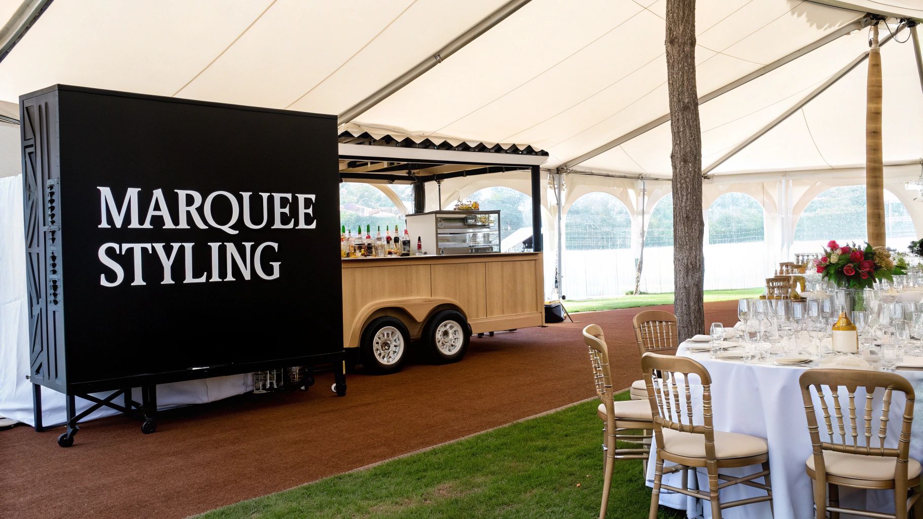 Inside a spacious marquee tent, a mobile bar and a beautifully set dining table with wooden chairs are ready for an event.