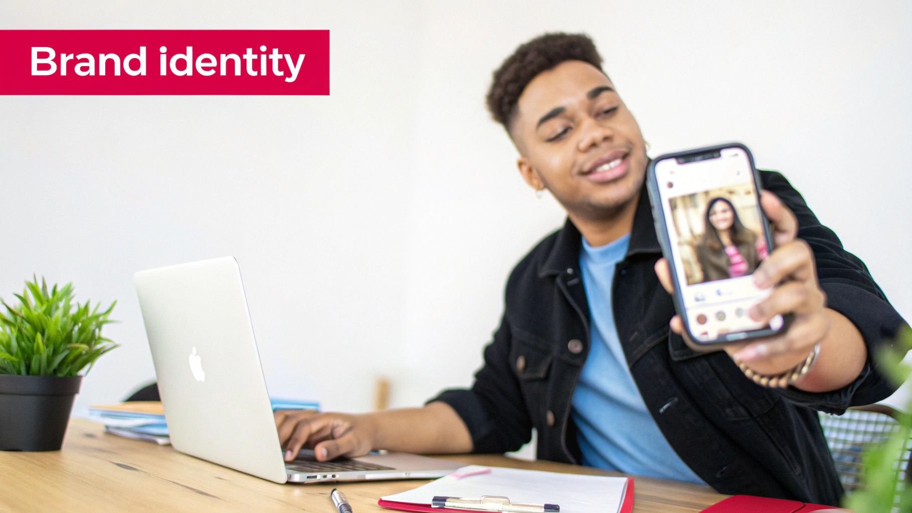 A smiling young man working on a laptop while showcasing a photo on his phone, related to brand identity.